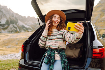 Naklejka premium Young woman with a phone enjoying the mountain landscape while sitting in the trunk of a car. Beautiful traveler blogging on a car adventure. Travel, freedom concept. Active lifestyle.