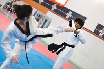 A female martial artist in a white gi and black belt delivers a high kick toward a male partner holding a target pad while both train on blue and red mats in an indoor dojo