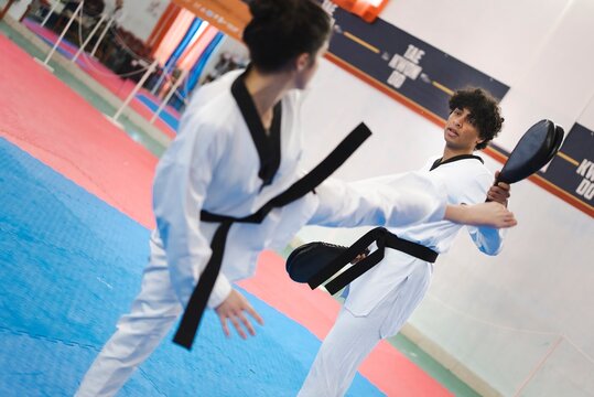 A female martial artist in a white gi and black belt delivers a high kick toward a male partner holding a target pad while both train on blue and red mats in an indoor dojo