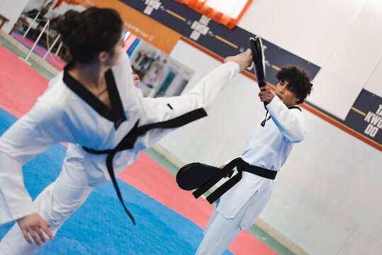 A female martial artist in a white gi and black belt delivers a high kick toward a male partner holding a target pad while both train on blue and red mats in an indoor dojo