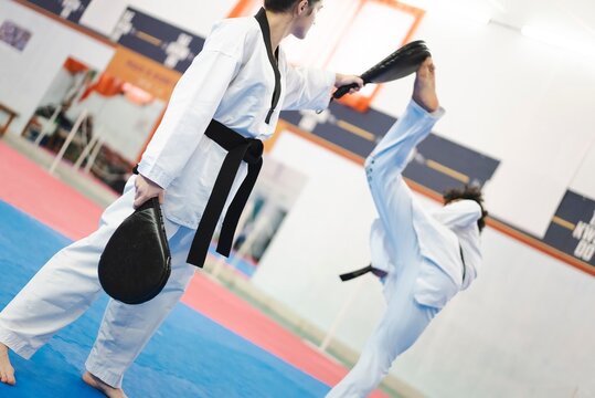 A martial artist in a white uniform and black belt extends a high leg kick toward a target pad held by a partner on a blue mat during intense indoor martial arts training