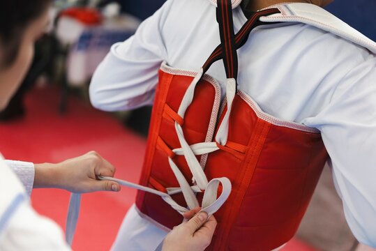 A martial artist adjusts and secures a red padded chest guard with white laces during training indoors while wearing a white uniform and black belt in a taekwondo dojo