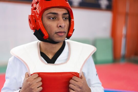 A male martial artist stands indoors on a mat dressed in white with a red protective helmet and chest guard while preparing to spar in a taekwondo training session
