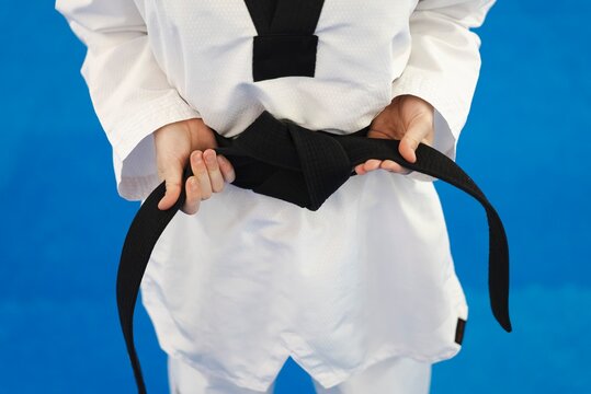 A close-up view of a martial artist's hands holding and tightening a black belt on their white uniform while standing on a blue training mat indoors during a taekwondo session