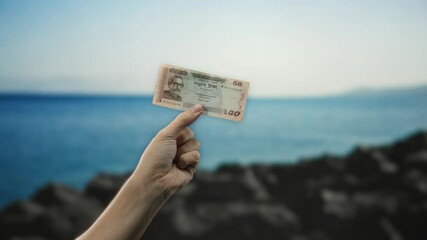 Man holds bangladesh banknote by seaside with ocean in background reflecting economic theme with outdoor and travel focus.