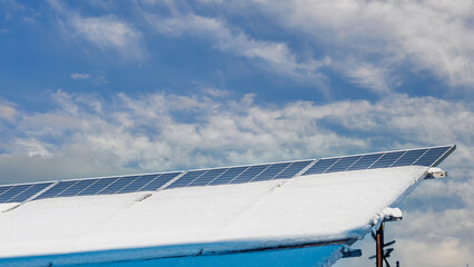 Solar panels on a roof covered with snow against a bright, cloudy sky in winter.