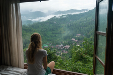 Person sitting on edge of bed looking at misty mountain landscape outside window during overcast day