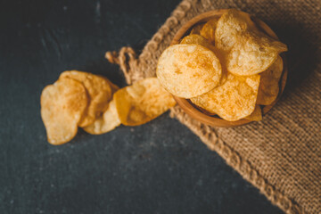 Crispy potato chips in a rustic wooden bowl on burlap fabric, with scattered chips on a dark...