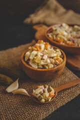 Close-up of traditional Olivier salad with peas, ham, potatoes, carrots, and eggs served in a rustic wooden bowl and spoon on burlap cloth with garlic and pickles on a dark background
