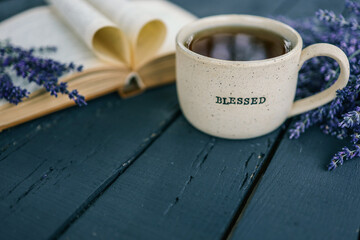 Ceramic cup with “BLESSED” text filled with tea, next to a heart-shaped open book and lavender flowers on a rustic blue wooden table, cozy and peaceful morning vibe