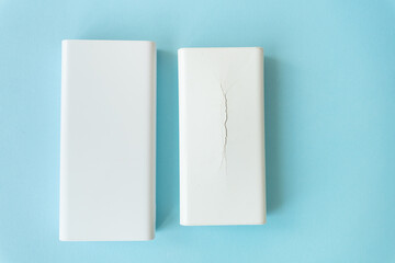Two white rectangular power banks, one undamaged and the other cracked, rest on a light blue...