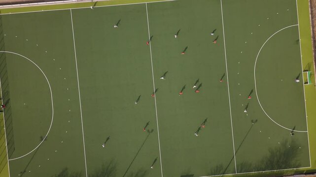 Angled drone shot of an outdoor hockey match on green turf with clear shadows and dynamic team formations.