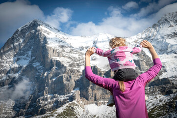 Two-year-old baby sitting on her mother's shoulders enjoying the view of the snow-capped mountains of the Swiss Alps