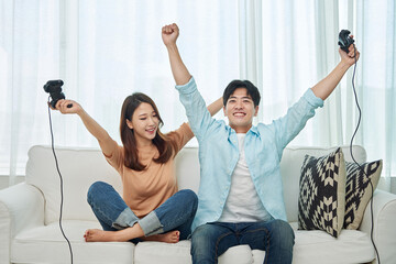 A young Asian man and woman are sitting on a living room sofa, holding game controllers, watching...