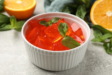 Delicious jelly cubes in bowl, mint and orange on grey table, closeup
