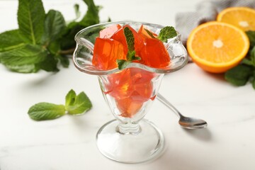 Delicious jelly cubes in dessert bowl, mint, spoon and orange on white marble table, closeup