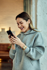 Fototapeta premium Laughing Woman Wearing Hoodie and Using Smartphone Indoors in Modern Home