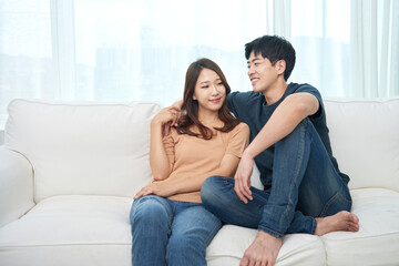 Young Asian man and woman sitting on a living room couch, affectionately putting their arms around each other's shoulders