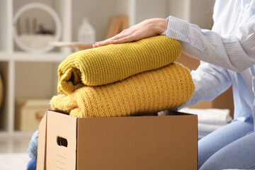 Young woman packing stack of clothes into cardboard box in bedroom, closeup