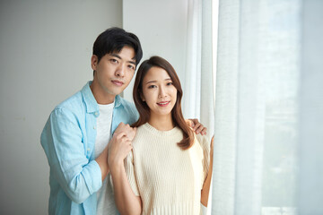 A young Asian man and a beautiful woman are conversing in a room with large windows adorned with white curtains, sipping warm drinks together.