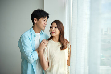 A young Asian man and a beautiful woman are conversing in a room with large windows adorned with...