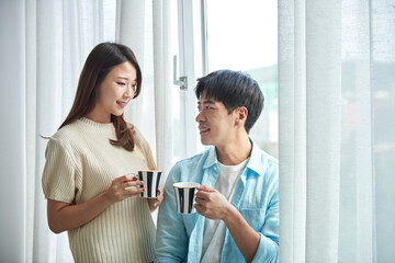 A young Asian man and a beautiful woman are conversing in a room with large windows adorned with...