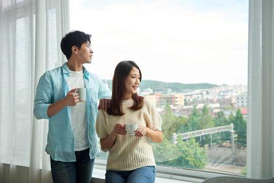 Young Asian couple enjoying the scenery through a large glass window, sipping coffee with happy expressions.