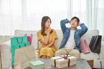 An Asian male and female couple is sitting on a living room sofa, regretting their excessive shopping.