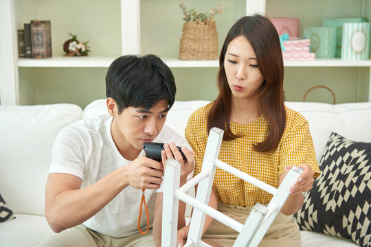 The newlywed couple sits on the living room sofa, assembling a chair with an electric drill