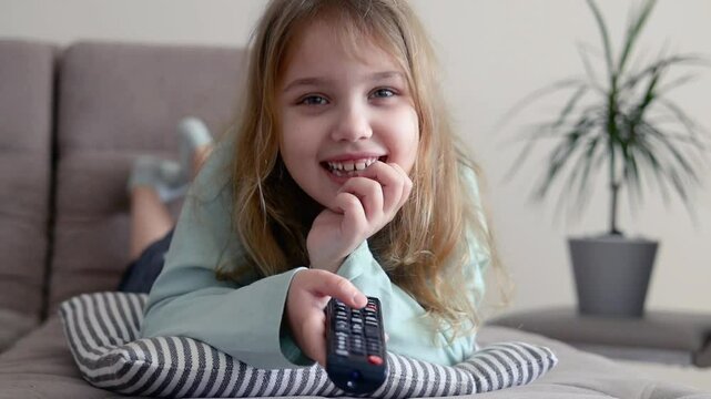 Caucasian child girl laying on sofa home and watching tv,using pilot,remote control.Happy kid indoors have a fun.Lesure concept.