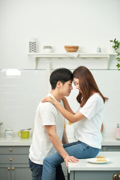 "A lovely couple in white cotton tees and jeans sits the woman on the kitchen table, engaging in affectionate skinship