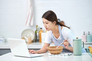 A young Asian woman cooks in the kitchen while checking her laptop