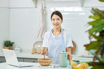 A young Asian woman cooks in the kitchen while checking her laptop