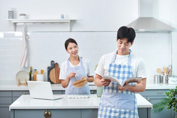 The newlywed couple in the kitchen, the young wife cooks while checking her laptop, as her apron-clad husband stands nearby holding a book, waiting for the meal