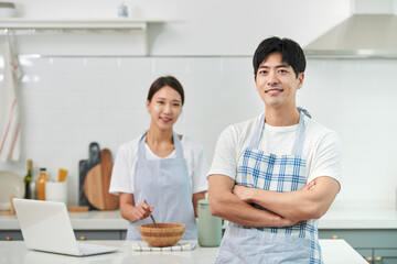 The newlywed couple is in the kitchen: the young wife prepares the meal while her husband stands nearby, smiling as he waits for the food