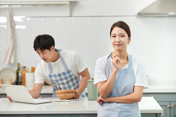 The newlyweds are in the kitchen: the husband is cooking while the wife stands nearby waiting for the meal to be ready
