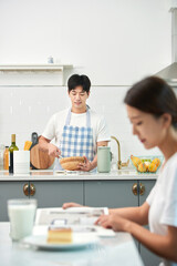 The young husband is cooking in the kitchen while his wife sits at the dining table reading a book