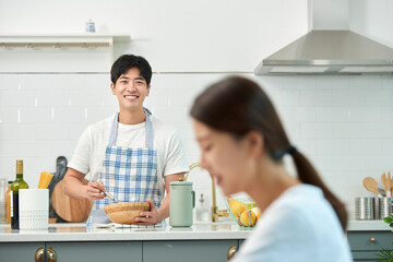 The young husband is cooking in the kitchen while his wife sits at the dining table reading a book