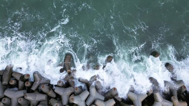 Aerial view of waves crashing against concrete tetrapods on a coastal barrier, creating dramatic white splashes and patterns