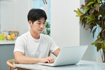 Young Asian man in a white T-shirt working on a laptop while sitting at a dining table chair in a living room with a kitchen