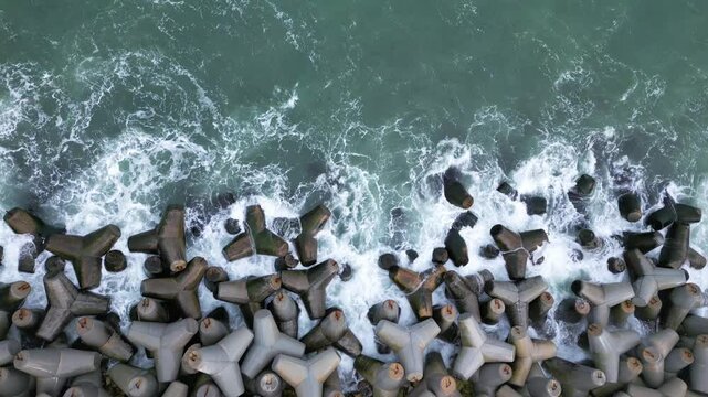 Aerial view of waves crashing against concrete tetrapods on a coastal barrier, creating dramatic white splashes and patterns