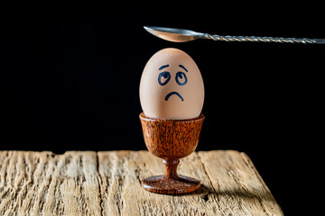 Metal spoon breaking boiled egg with sad face in egg cup on wooden table, closeup. Scared egg looking at spoon