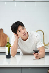 A young Asian man at the kitchen table holding a wine bottle and a wine glass, struggling with a...