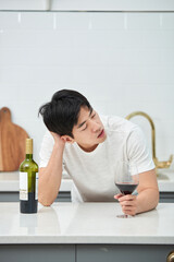 A young Asian man at the kitchen table holding a wine bottle and a wine glass, struggling with a...