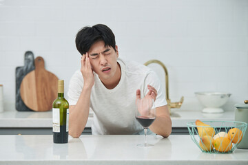 A young Asian man at the kitchen table holding a wine bottle and a wine glass, struggling with a...