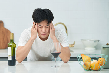 A young Asian man at the kitchen table holding a wine bottle and a wine glass, struggling with a...