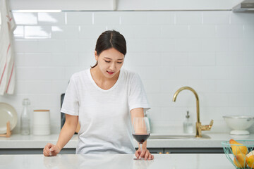 A young Asian woman at the kitchen table holding a wine glass, struggling with a hangover from the...