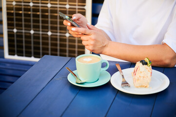 Young woman charging her smartphone from solar panel while relaxing. Pretty girl drinking coffee and checking her phone next to solar battery. Conscious female choosing sustainable lifestyle.