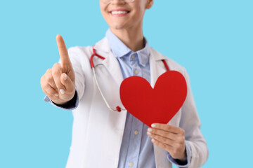 Female doctor with paper heart pointing at something on blue background, closeup. Blood donation...