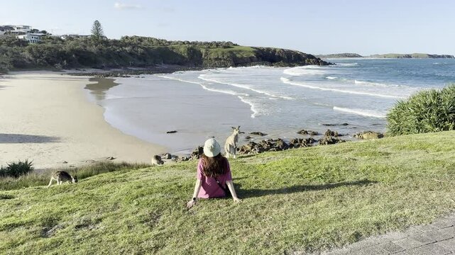 A happy girl wearing a hat sits on the grass surrounded by eastern gray kangaroos feeding on a hill overlooking the stunning Shelly Beach. Look at me now headland walk, New South Wales, Australia